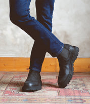 A man in jeans and Bed Stu Grit black boots standing on a rug.
