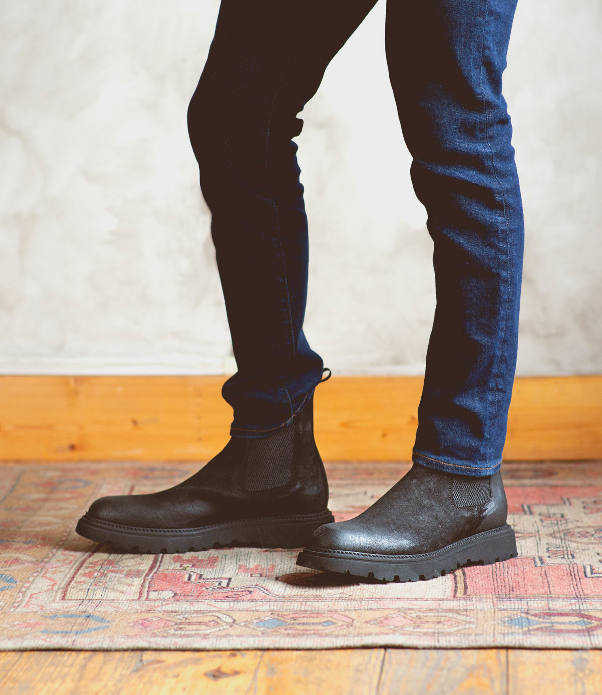 A man exuding casual elegance, standing on a rug in Bed Stu Grit Chelsea boots of enduring quality.