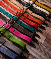 Bed Stu Crossbody Straps in various colors and adjustable leather designs with metal clasps are displayed diagonally on a wooden surface.