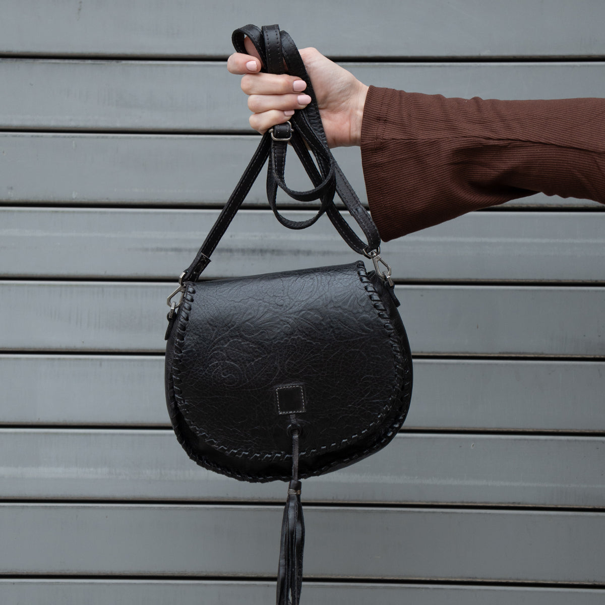 A person in a brown long-sleeve top holds up the Bed Stu Half Moon EM black saddlebag crossbody purse with a leather tassel, standing in front of a gray metal shutter.
