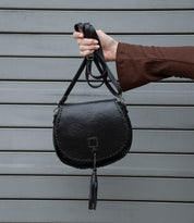 A person in a brown long-sleeve top holds up the Bed Stu Half Moon EM black saddlebag crossbody purse with a leather tassel, standing in front of a gray metal shutter.