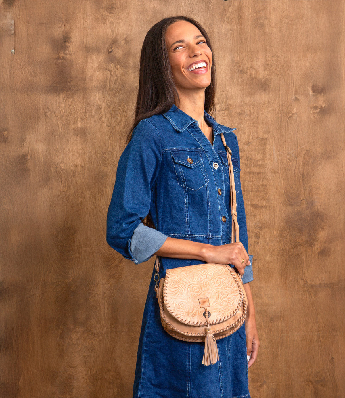 A woman in a denim dress stands smiling by a wooden wall, carrying the Bed Stu Half Moon EM tan saddlebag crossbody purse with an embossed foliage pattern over her shoulder.