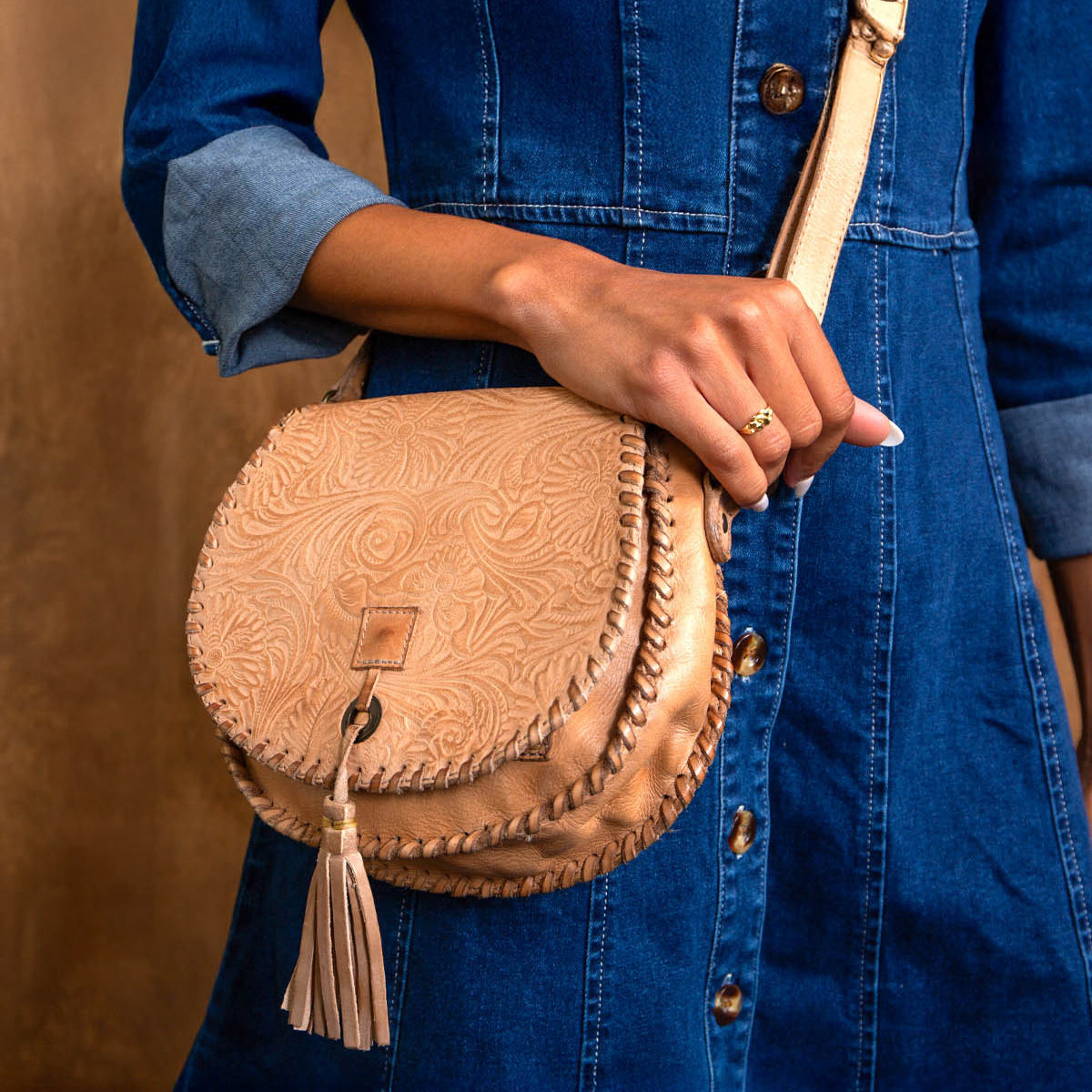 A person in a blue denim dress holds the Bed Stu Half Moon EM saddlebag crossbody, featuring an embossed foliage pattern and leather tassel detail.