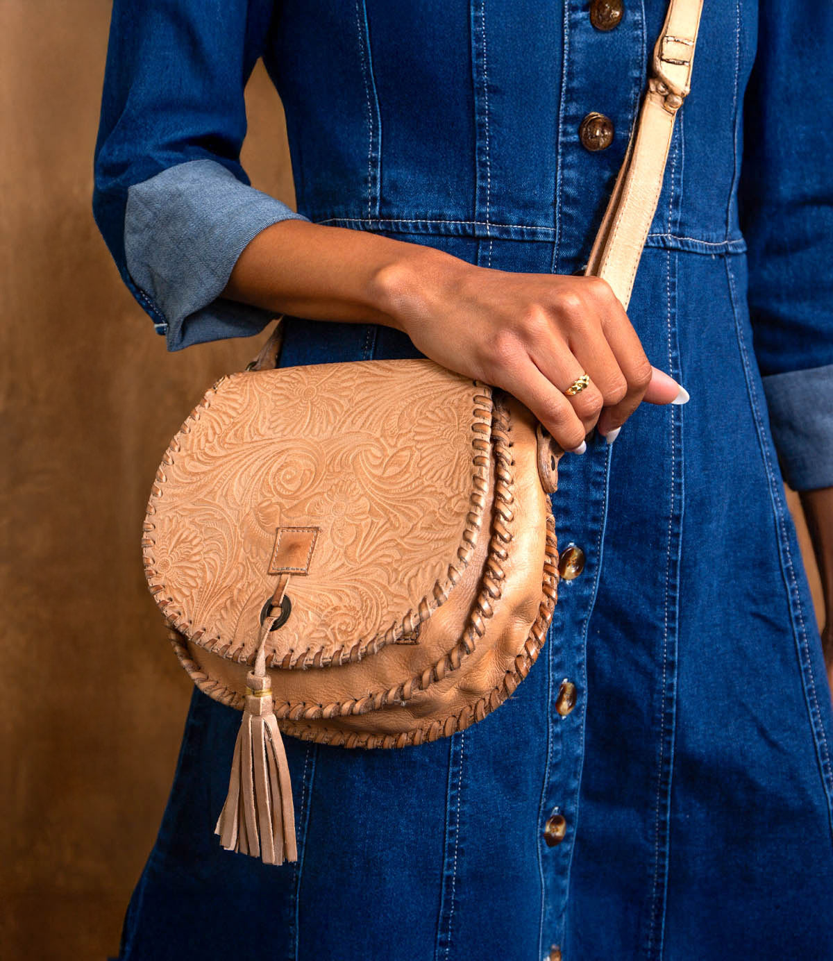 A person in a blue denim dress holds the Bed Stu Half Moon EM saddlebag crossbody, featuring an embossed foliage pattern and leather tassel detail.