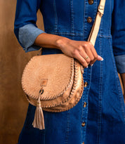 A person in a blue denim dress holds the Bed Stu Half Moon EM saddlebag crossbody, featuring an embossed foliage pattern and leather tassel detail.