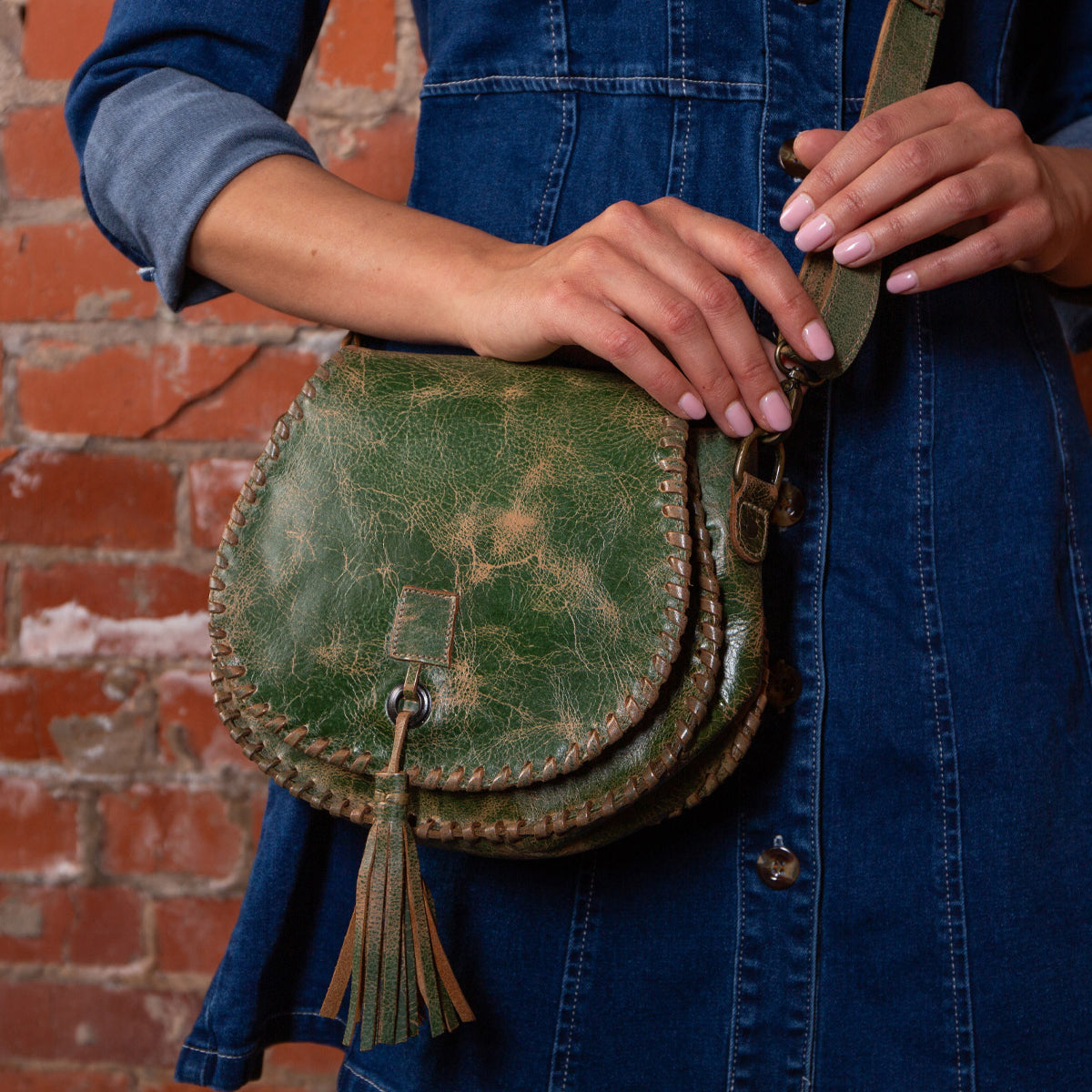 A person in a denim dress holds the Bed Stu Half Moon green crossbody purse with adjustable strap and leather tassel, standing by a red brick wall.