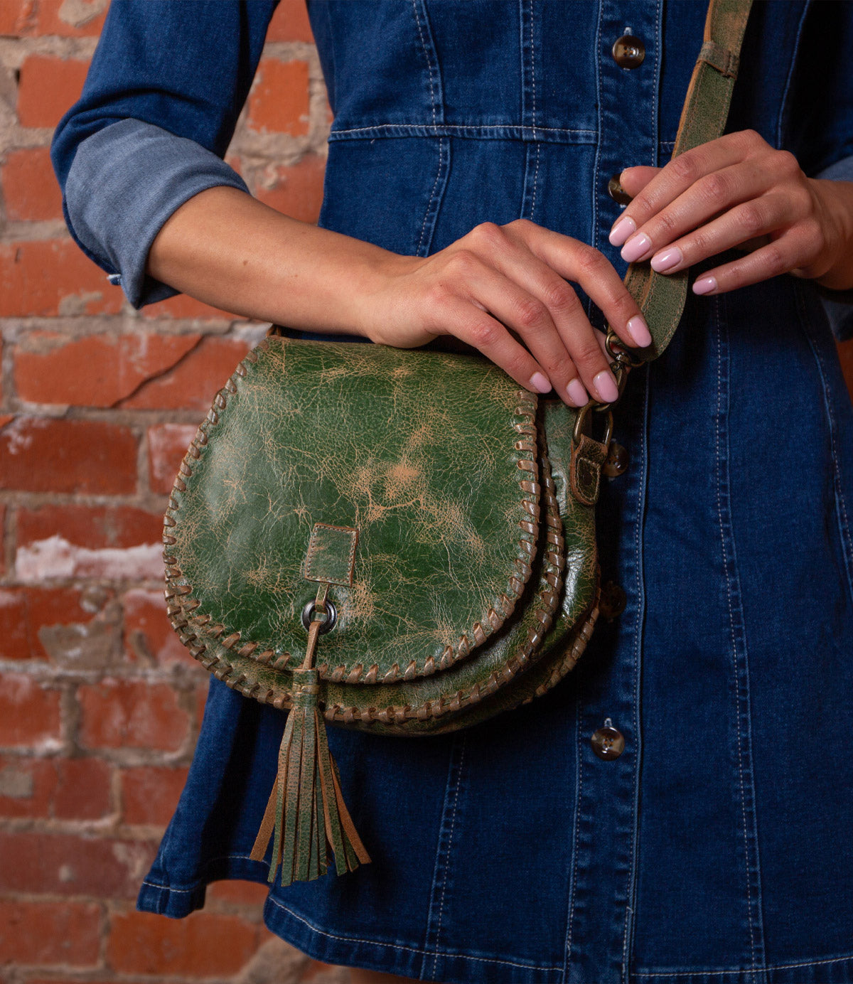 A person in a denim dress holds the Bed Stu Half Moon green crossbody purse with adjustable strap and leather tassel, standing by a red brick wall.