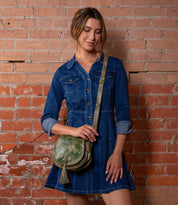 A woman stands before a brick wall wearing a blue denim dress with rolled sleeves and carrying the Bed Stu Half Moon crossbody purse in green, which features an adjustable strap.
