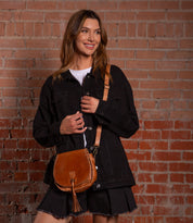 A woman in a black denim jacket and black shorts stands against a brick wall, smiling as she holds the Bed Stu Half Moon brown crossbody purse with an adjustable strap.