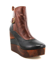 The Bed Stu Harmonious is a brown and black leather Platform Wedge Ankle Boot with a sculptural wood wedge sole, brogue-style perforations, and a decorative side buckle.