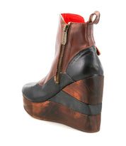 The Bed Stu Harmonious is a brown and black platform wedge ankle boot featuring a side zipper, brogue-style perforations, and a thick, layered wood-look sole.