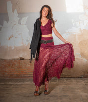 A woman stands indoors in a burgundy lace dress, holding a black jacket and lifting her skirt, wearing Bed Stu’s Harmonious sculptural wood wedge platform boots amid leather details, worn walls, and exposed brick.