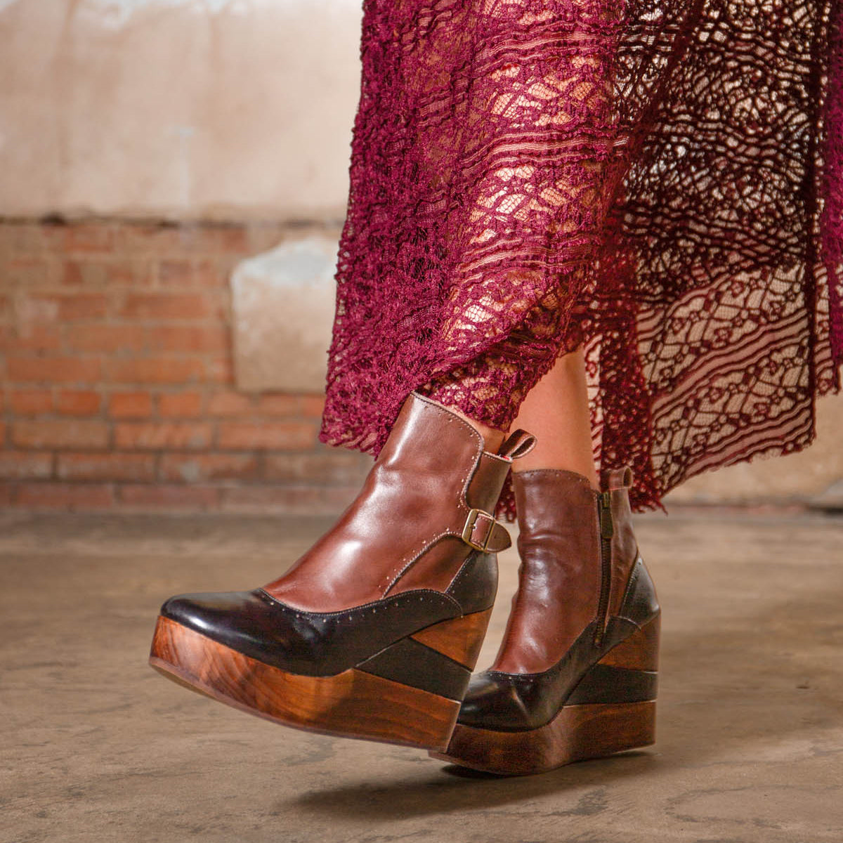 A person wearing Bed Stu's Harmonious brown and black Platform Wedge Ankle Boots with sculptural wood wedge and buckle, paired with a burgundy lace skirt, stands on a concrete floor in front of a brick wall.