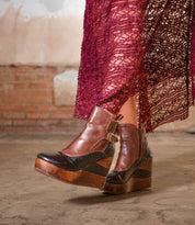 A person wearing Bed Stu's Harmonious brown and black Platform Wedge Ankle Boots with sculptural wood wedge and buckle, paired with a burgundy lace skirt, stands on a concrete floor in front of a brick wall.