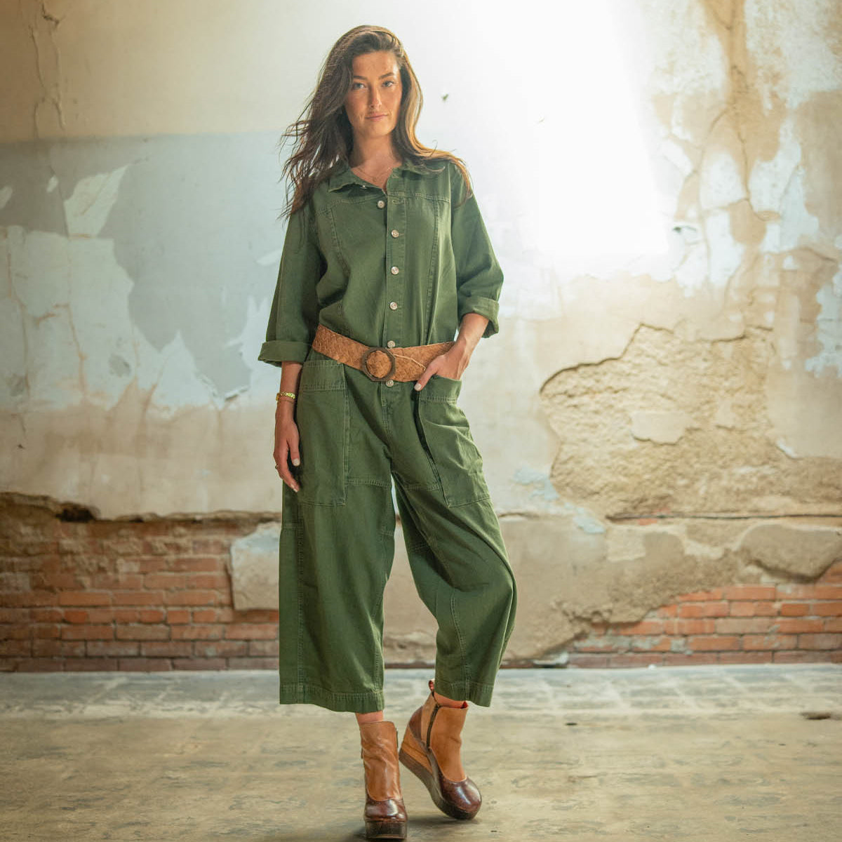 A woman stands indoors wearing the Bed Stu Harmonious sculptural wood wedge platform shoes paired with a green jumpsuit and brown belt, set against a worn wall with exposed brick and peeling paint.