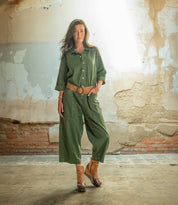 A woman stands indoors wearing the Bed Stu Harmonious sculptural wood wedge platform shoes paired with a green jumpsuit and brown belt, set against a worn wall with exposed brick and peeling paint.