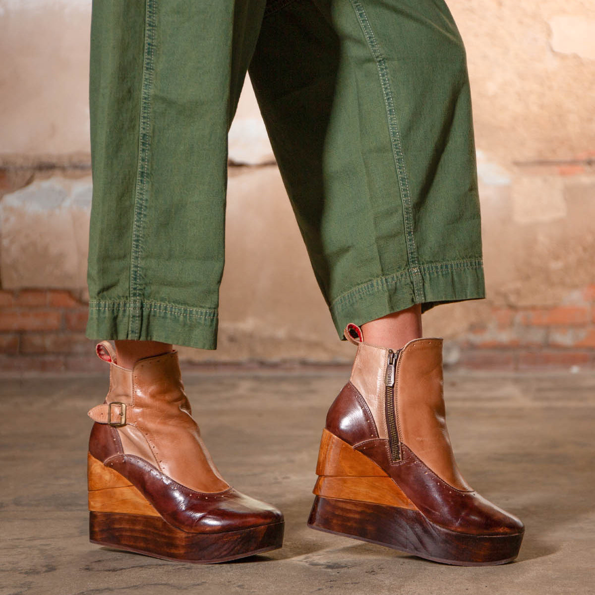 A person in green pants wears Bed Stu’s Harmonious Platform Wedge Ankle Boots, featuring sculptural wood wedges, zippers, and buckles, while standing on a concrete floor with a brick and plaster wall behind them.