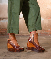 A person in green pants wears Bed Stu’s Harmonious Platform Wedge Ankle Boots, featuring sculptural wood wedges, zippers, and buckles, while standing on a concrete floor with a brick and plaster wall behind them.