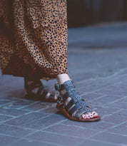 A person wearing the Bed Stu Hera gladiator sandals with a stylish leather outsole and a leopard print dress walks along a paved surface.