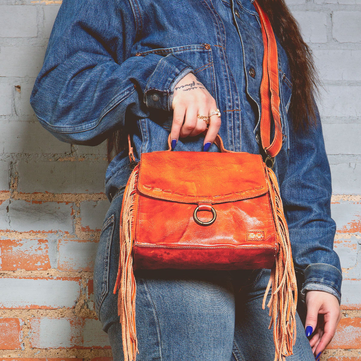 A person in a denim jacket holds a Bed Stu crossbody bag crafted from orange leather, featuring fringe accents and an adjustable strap.
