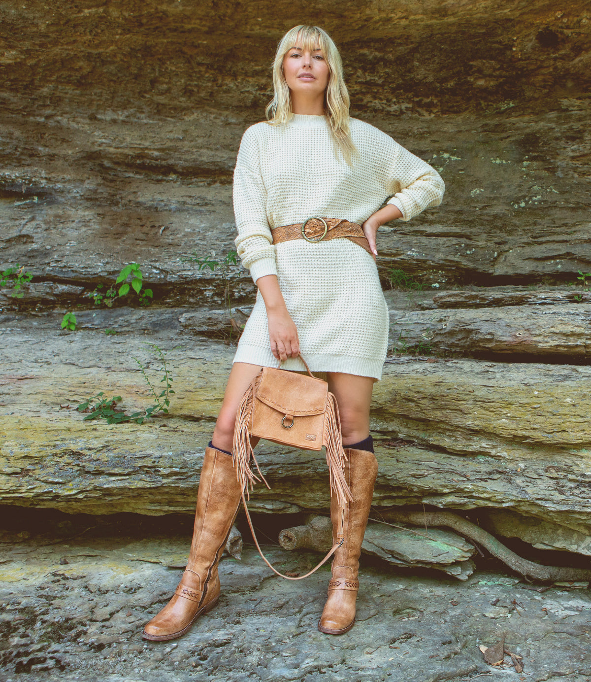 A blonde woman in a cream sweater dress and cowboy boots, carrying a Bed Stu Hidden leather crossbody bag with adjustable and removable crossbody strap.