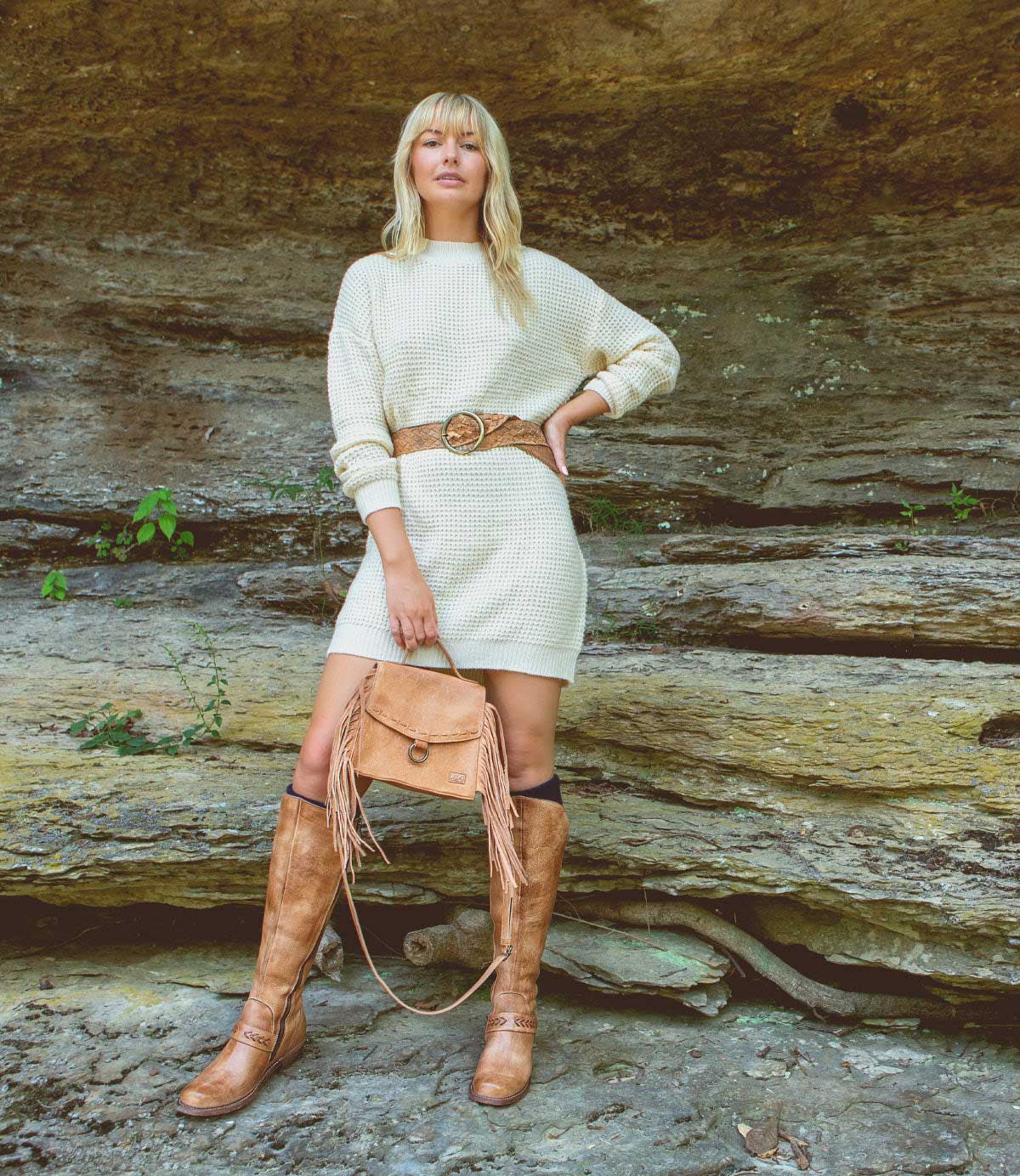 A blonde woman in a cream sweater dress and cowboy boots, carrying a Bed Stu Hidden leather crossbody bag with adjustable and removable crossbody strap.