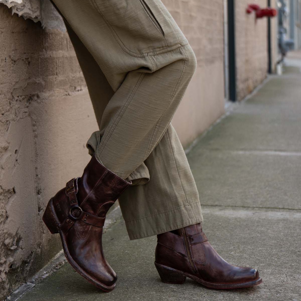 A person wearing olive green cargo pants and Bed Stu's Homage brown leather harness boots with Vibram panels leans against a textured beige wall on a concrete sidewalk.