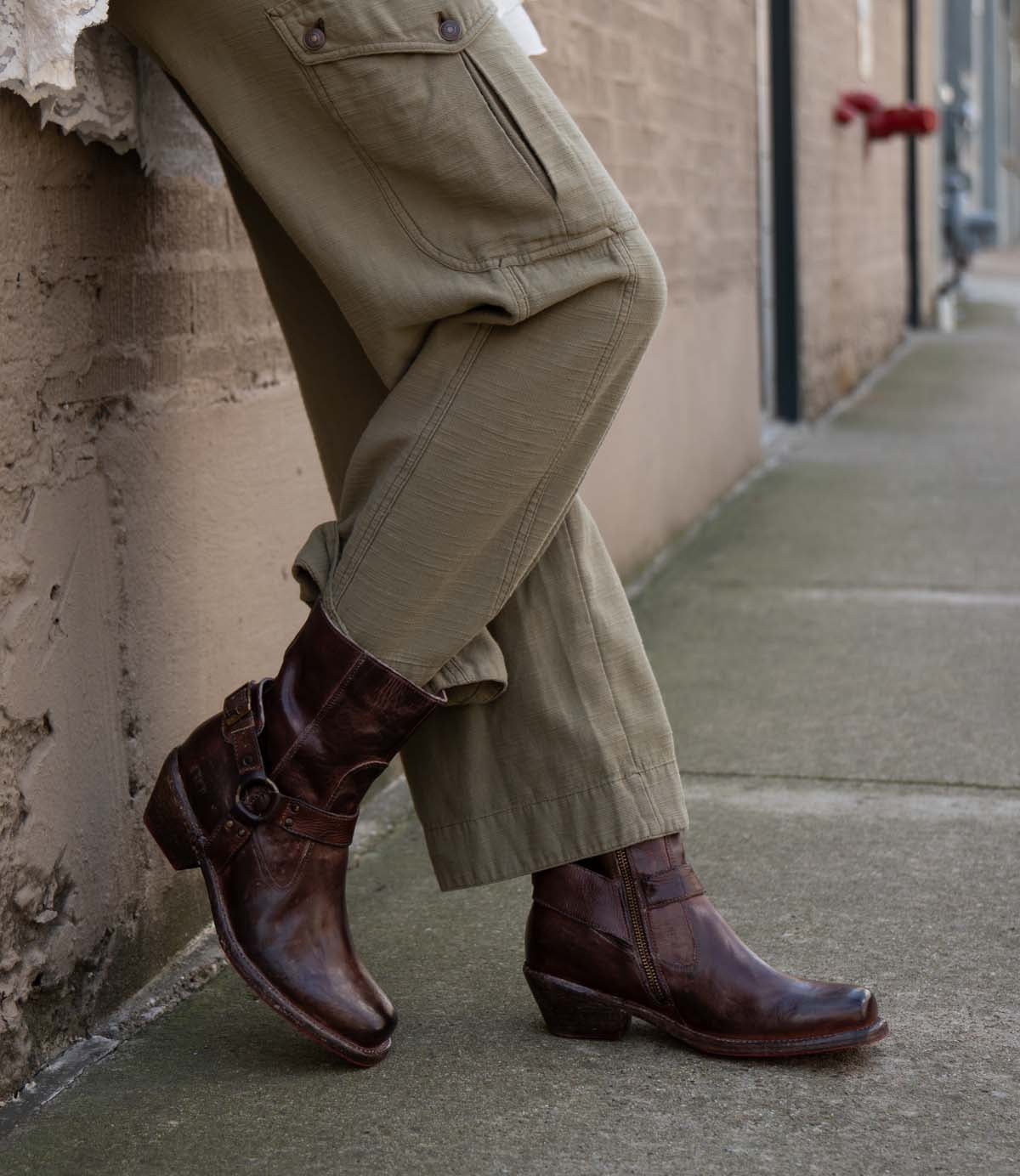 A person wearing olive green cargo pants and Bed Stu's Homage brown leather harness boots with Vibram panels leans against a textured beige wall on a concrete sidewalk.