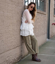 A woman in sunglasses leans against a brick wall, wearing a white lace top, olive green pants, and Bed Stu Homage brown leather harness boots.
