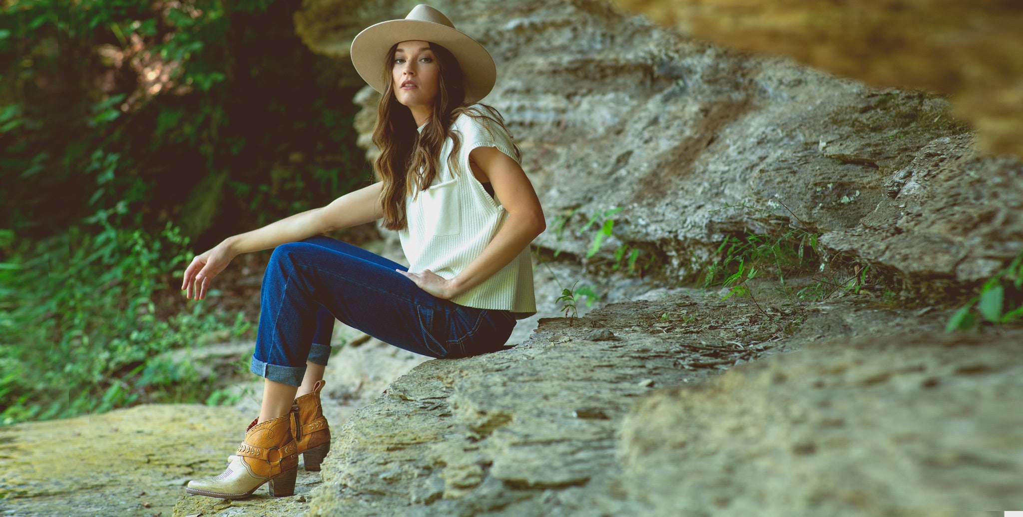 Woman in hat, cream top & dark denim sitting on a rock ledge wearing short leather western booties
