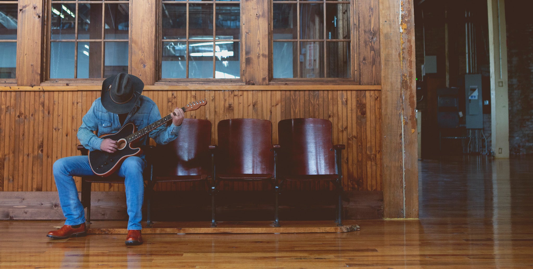 Man sitting on wooden stadium seats in wood building, wearing all denim & a cowboy hat, strumming a guitar