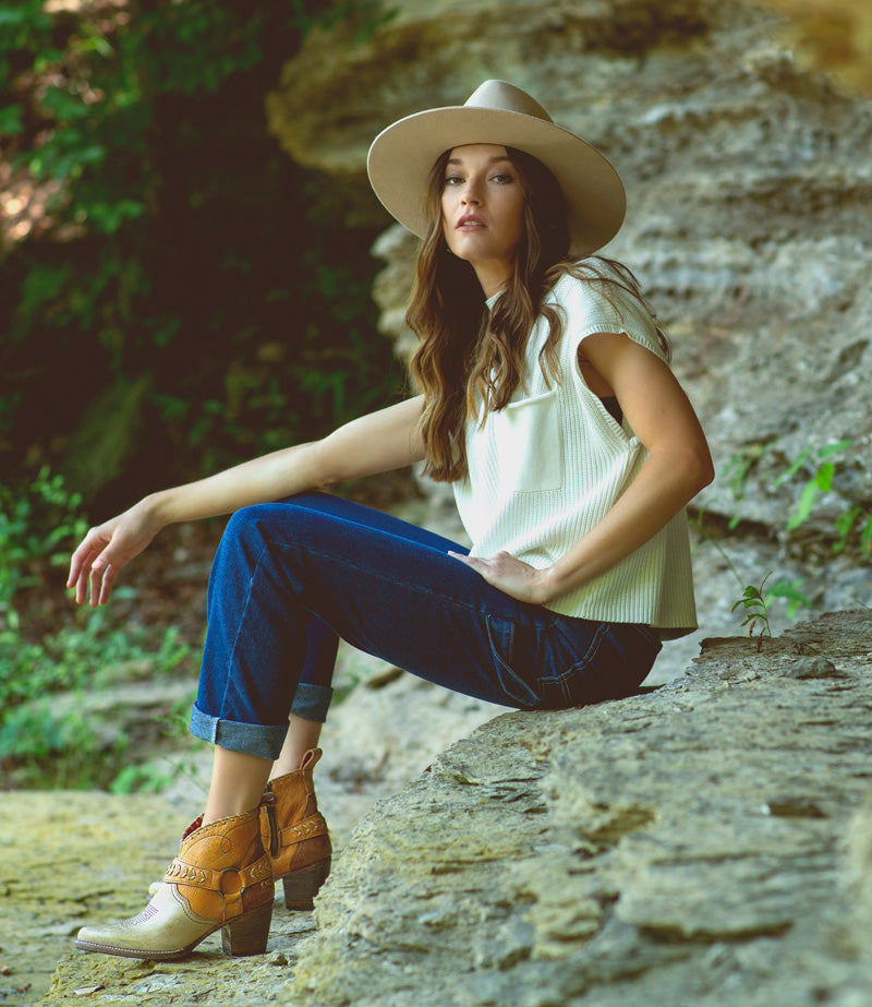 Woman in hat, cream top & dark denim sitting on a rock ledge wearing short leather western booties