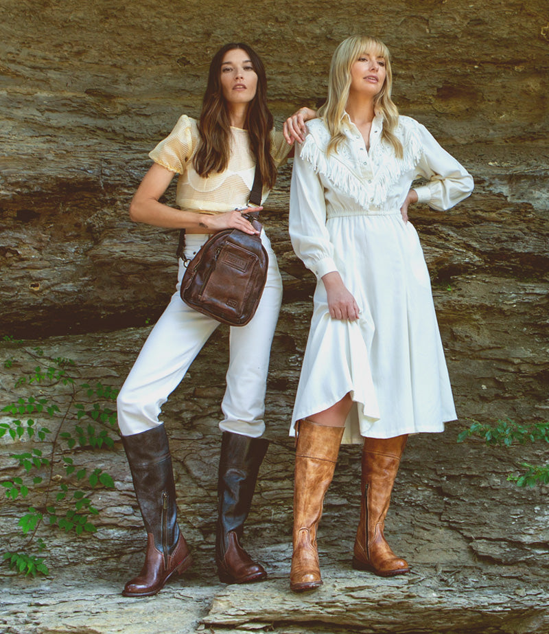 Two women in neutral outfits, standing on a rock ledge wearing tall leather boots 