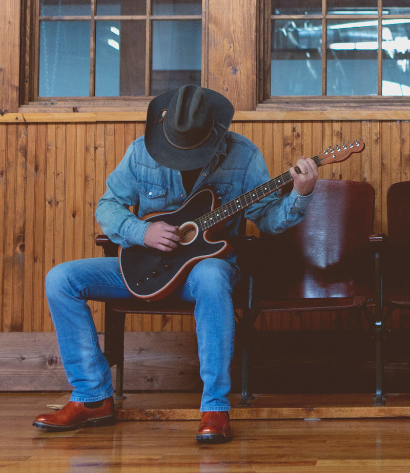 Man sitting on wooden stadium seats in wood building, wearing all denim & a cowboy hat, strumming a guitar