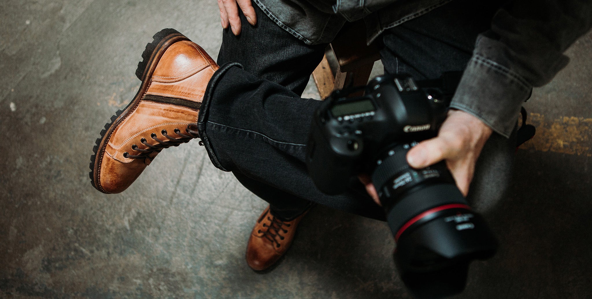 Top shot of mans hands holding camera over his crossed legs wearing tan leather boots.