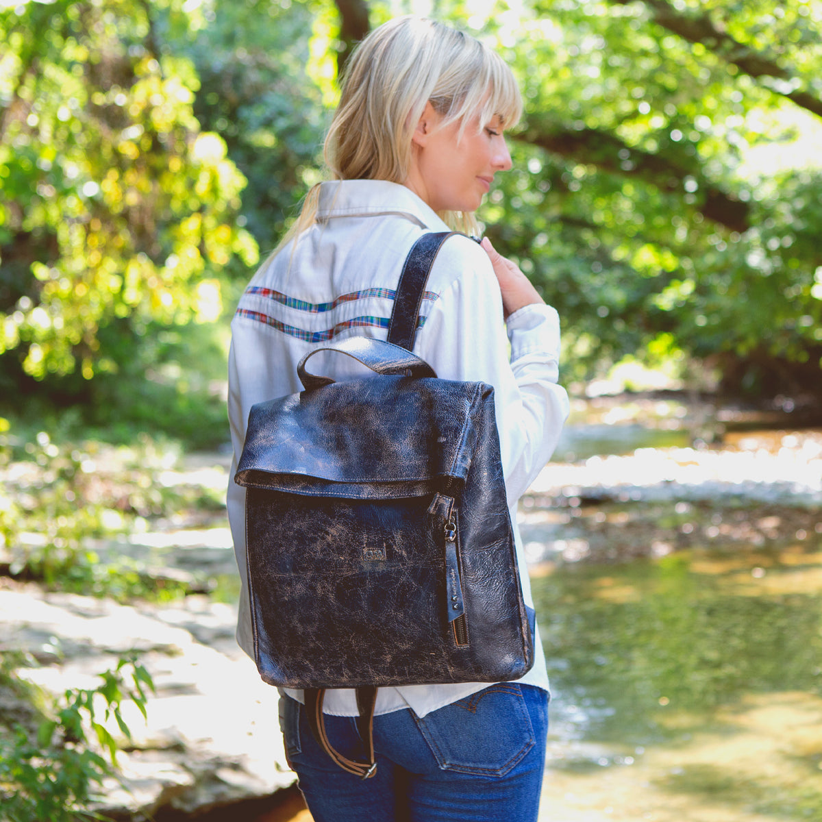 A woman standing by a river with a Bed Stu leather backpack named Howie.