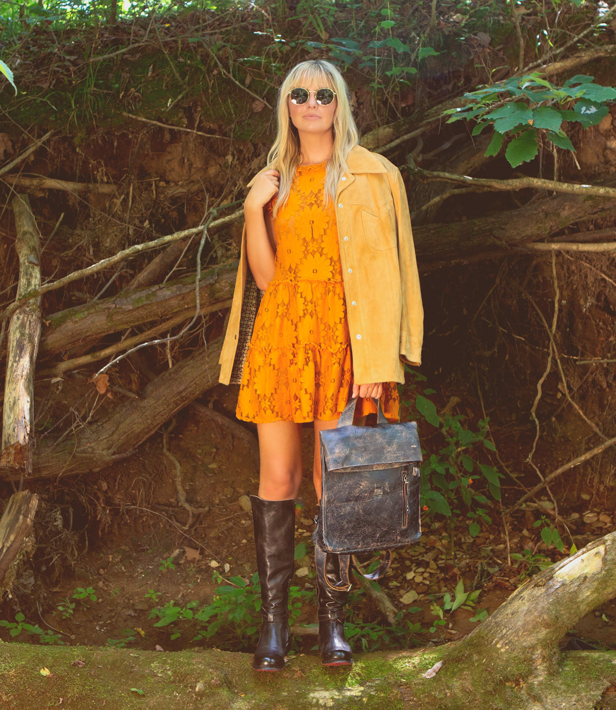 A blonde woman in a yellow dress standing in the woods holding a Bed Stu Howie leather bag.