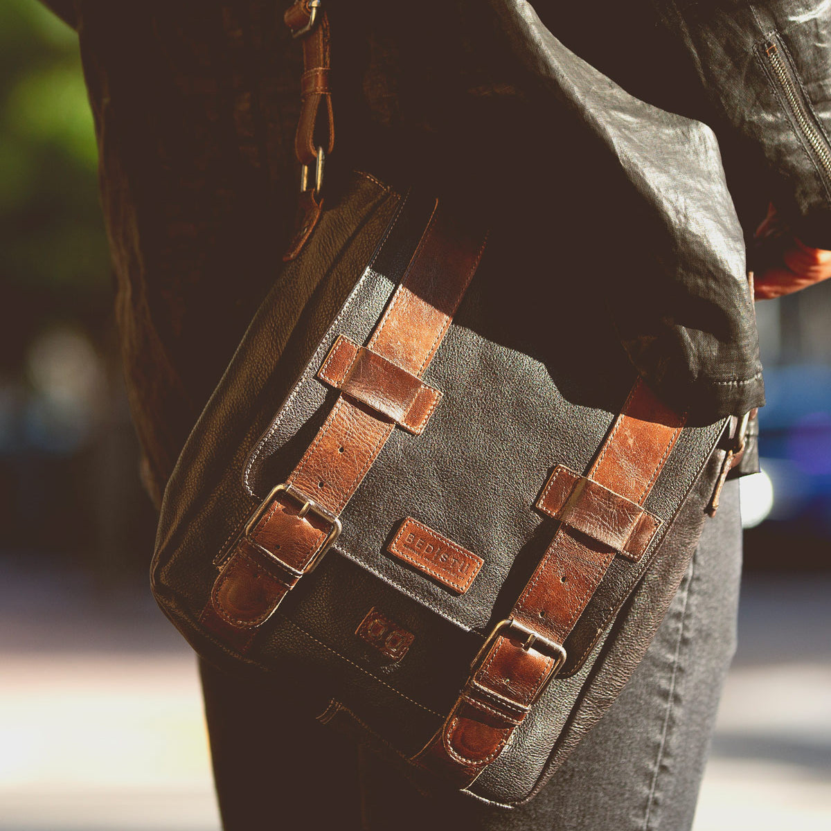 Close-up of a person carrying the versatile Hue brown leather messenger bag by Bed Stu, featuring two buckle straps and a small label on the front. The individual, dressed in a black jacket and gray pants, showcases the adjustable strap of this stylish dual-gender accessory.