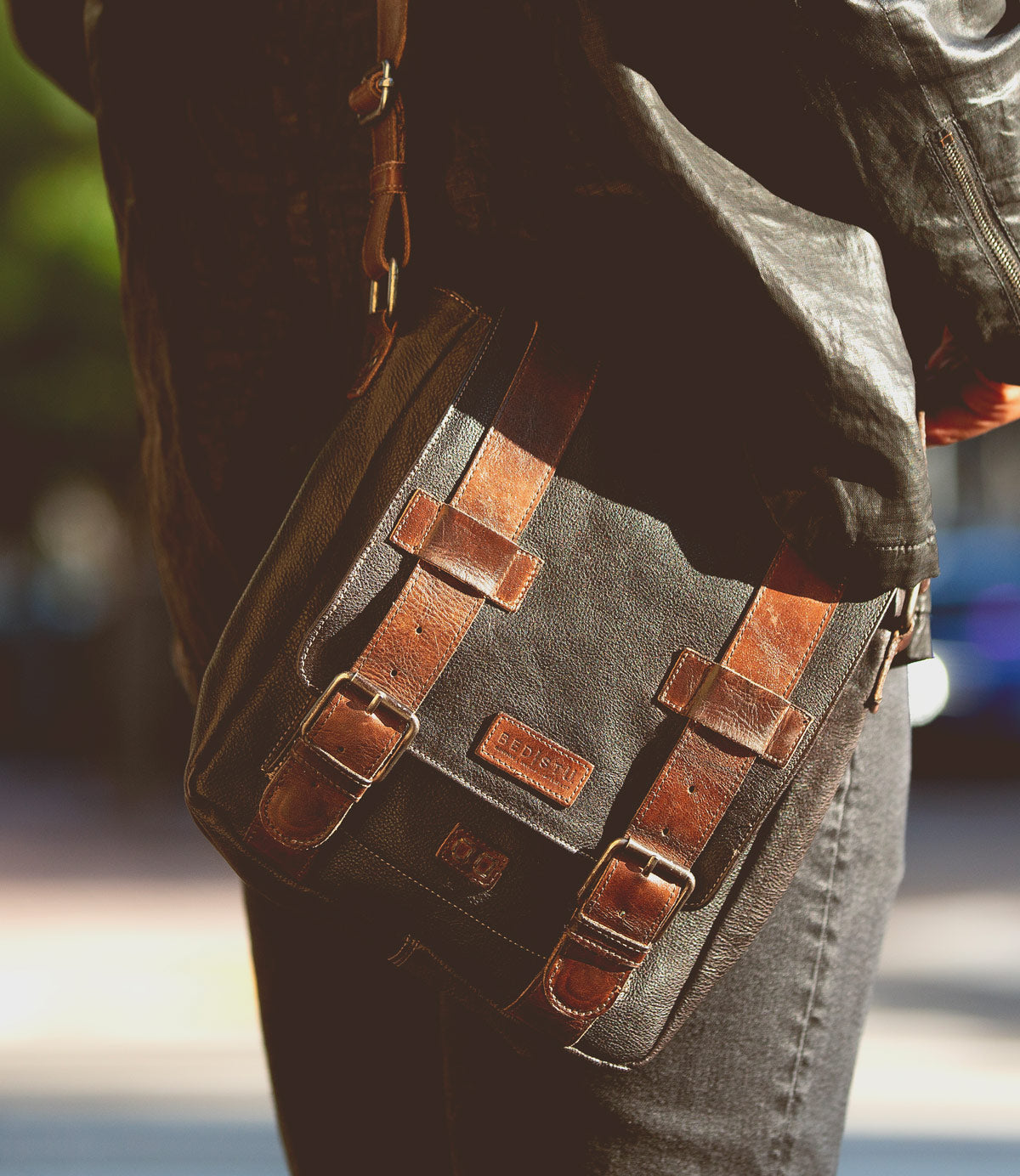 Close-up of a person carrying the versatile Hue brown leather messenger bag by Bed Stu, featuring two buckle straps and a small label on the front. The individual, dressed in a black jacket and gray pants, showcases the adjustable strap of this stylish dual-gender accessory.