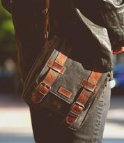 Close-up of a person carrying the versatile Hue brown leather messenger bag by Bed Stu, featuring two buckle straps and a small label on the front. The individual, dressed in a black jacket and gray pants, showcases the adjustable strap of this stylish dual-gender accessory.