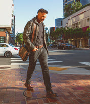 A man in dark clothing and a leather jacket, carrying the Hue bag by Bed Stu with an adjustable strap, walks across a brick sidewalk at an urban street intersection on a sunny day.