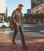 A man in dark clothing and a leather jacket, carrying the Hue bag by Bed Stu with an adjustable strap, walks across a brick sidewalk at an urban street intersection on a sunny day.