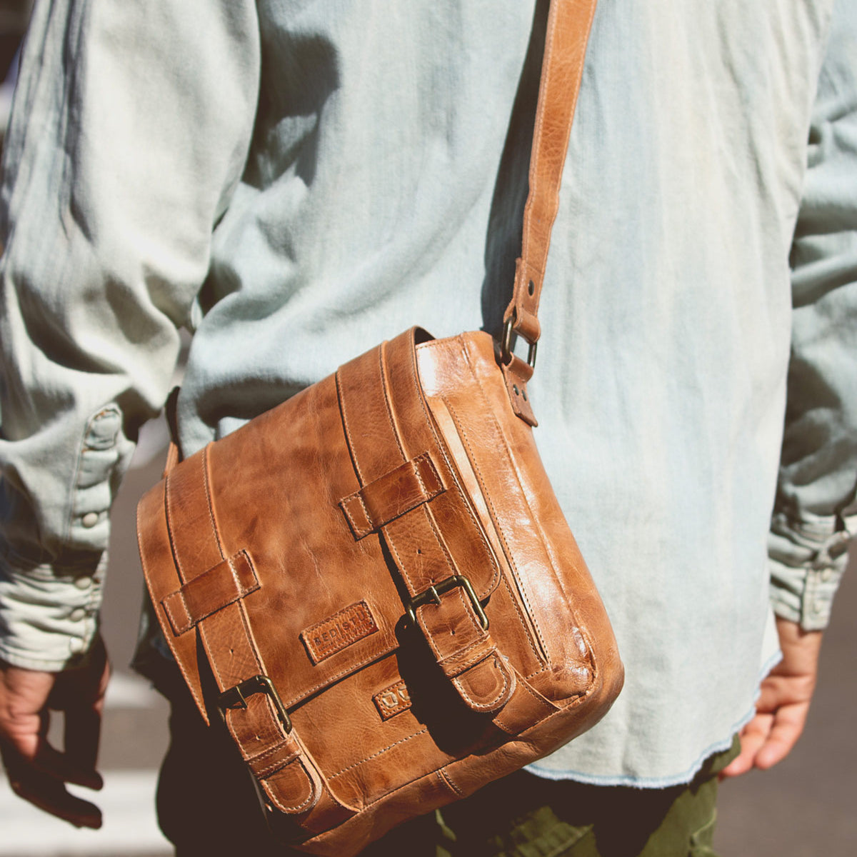 A person wearing a light green Hue shirt and carrying a tan Bed Stu leather messenger bag with an adjustable strap stands outdoors.