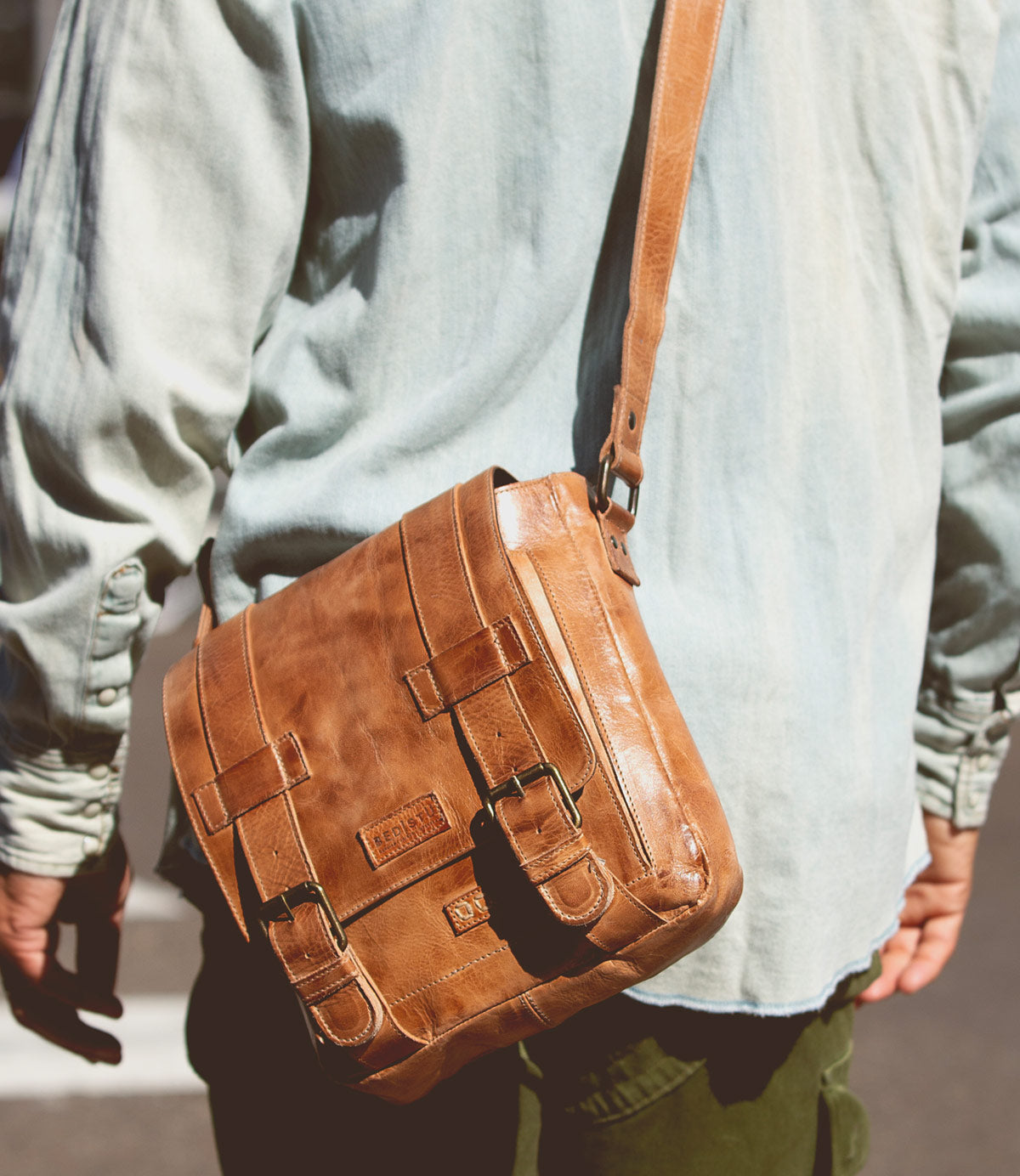A person wearing a light green Hue shirt and carrying a tan Bed Stu leather messenger bag with an adjustable strap stands outdoors.