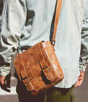 A person wearing a light green Hue shirt and carrying a tan Bed Stu leather messenger bag with an adjustable strap stands outdoors.