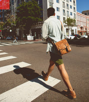 A person wearing a light shirt, green shorts, and carrying the versatile Bed Stu Hue messenger bag with an adjustable strap crosses a city street at a crosswalk on a sunny day.