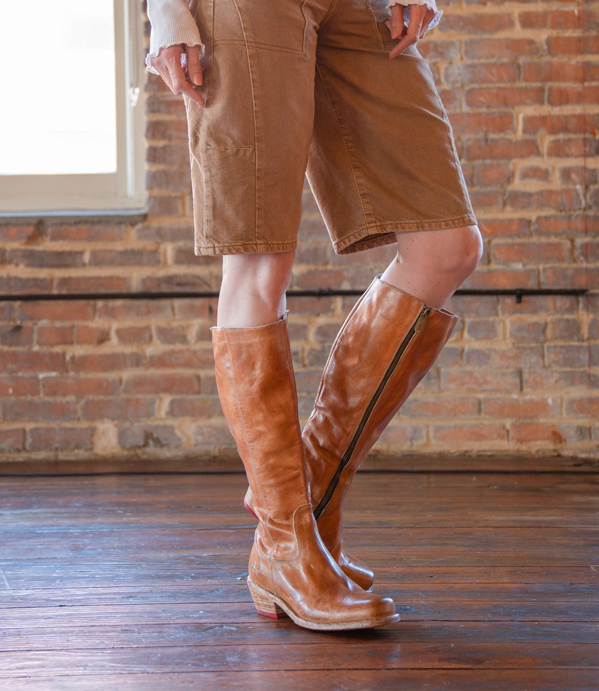 Wearing the Bed Stu Humble brown square toe boots with zippers and tan knee-length shorts on a wooden floor by a brick wall—these stylish boots are the perfect choice for your fall and winter look.