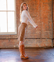 A woman stands indoors in profile, wearing a white top, tan skirt, and Bed Stu's Humble brown leather boots—perfect fall-winter footwear—with a brick wall and large window behind her.