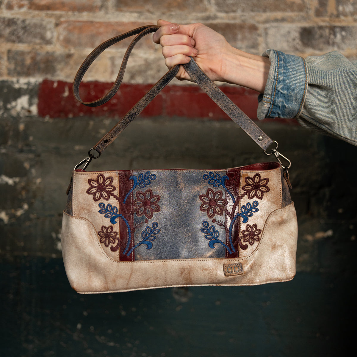 A hand holds the Bed Stu Hyperbolic, a handcrafted leather shoulder bag featuring blue and red floral embroidery, set against a distressed brick and painted wall backdrop.