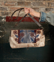 A hand holds the Bed Stu Hyperbolic, a handcrafted leather shoulder bag featuring blue and red floral embroidery, set against a distressed brick and painted wall backdrop.